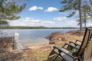 A wooden dock leading to a calm lake with chairs on the shore.