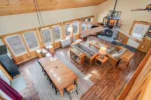 A spacious room with a wooden floor, a fireplace, a dining table, and large windows. - Lake Front Cabin on Desirable Highland Lake (Bridgton)