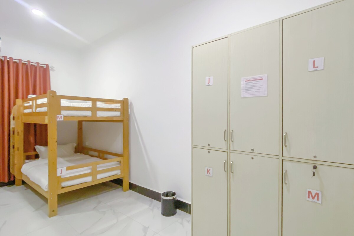 A bunk bed with a wooden frame and white bedding, a metal wardrobe with multiple compartments, and a trash bin on the floor.