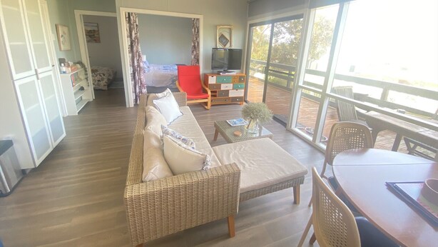 A spacious living area with a sofa, a dining table, and a TV. There is a balcony with a wooden deck and a view of the outdoors. - Straight to the Point - Sea views from balcony, dog friendly! (Pine Point)