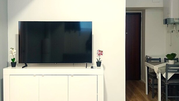 A modern living room with a flat-screen TV mounted on a white cabinet, a dining area with a table and chairs, and a kitchen with an air conditioning unit. - Mayflowers Apartment  - Biancospini (Milan)
