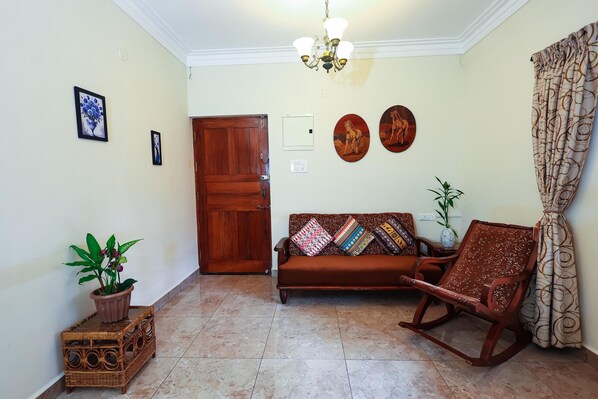 A living room with a wooden door, a brown sofa, a rocking chair, a potted plant, and a wall-mounted light fixture. - Vacation Home Stay Goa (Candolim)