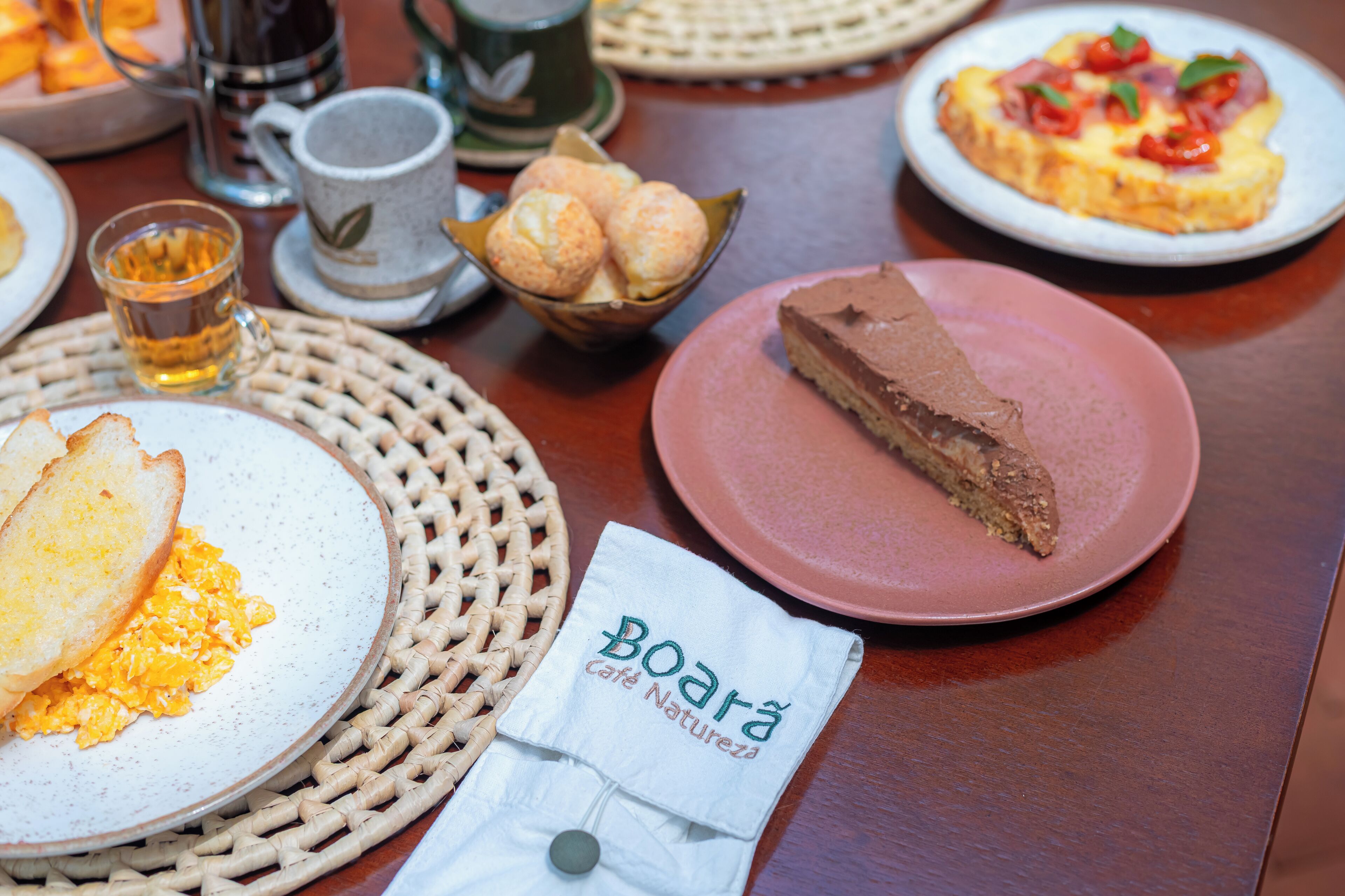 Uma mesa com itens de café da manhã, incluindo torradas, ovos mexidos, uma fatia de bolo de chocolate e um prato de pizza.