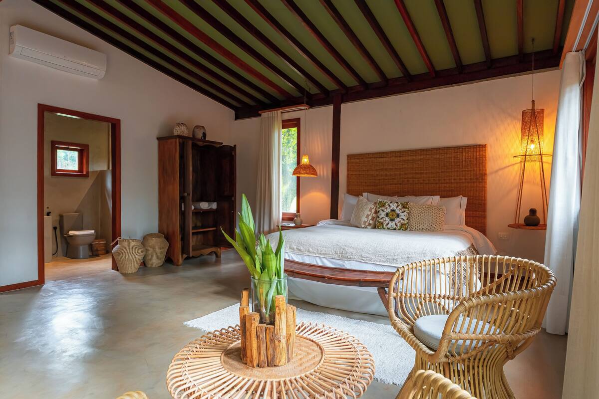 A bedroom with a large bed, a wicker chair, a bedside table, and a window with curtains.