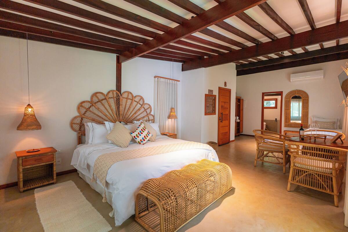 A bedroom with a large bed, a wooden bench, wicker chairs, a wooden table, and a hanging light fixture.
