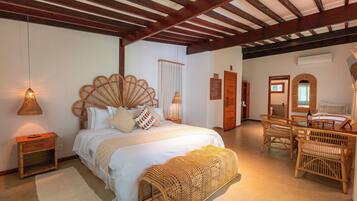 A bedroom with a large bed, a wooden bench, wicker chairs, a wooden table, and a hanging light fixture.