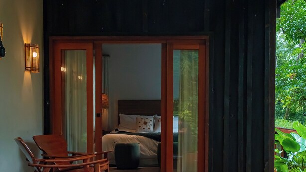 A wooden deck with a hot tub, a wooden rocking chair, and a bedroom with a bed and curtains. - Boarã Lodge Natureza (Pirenópolis)