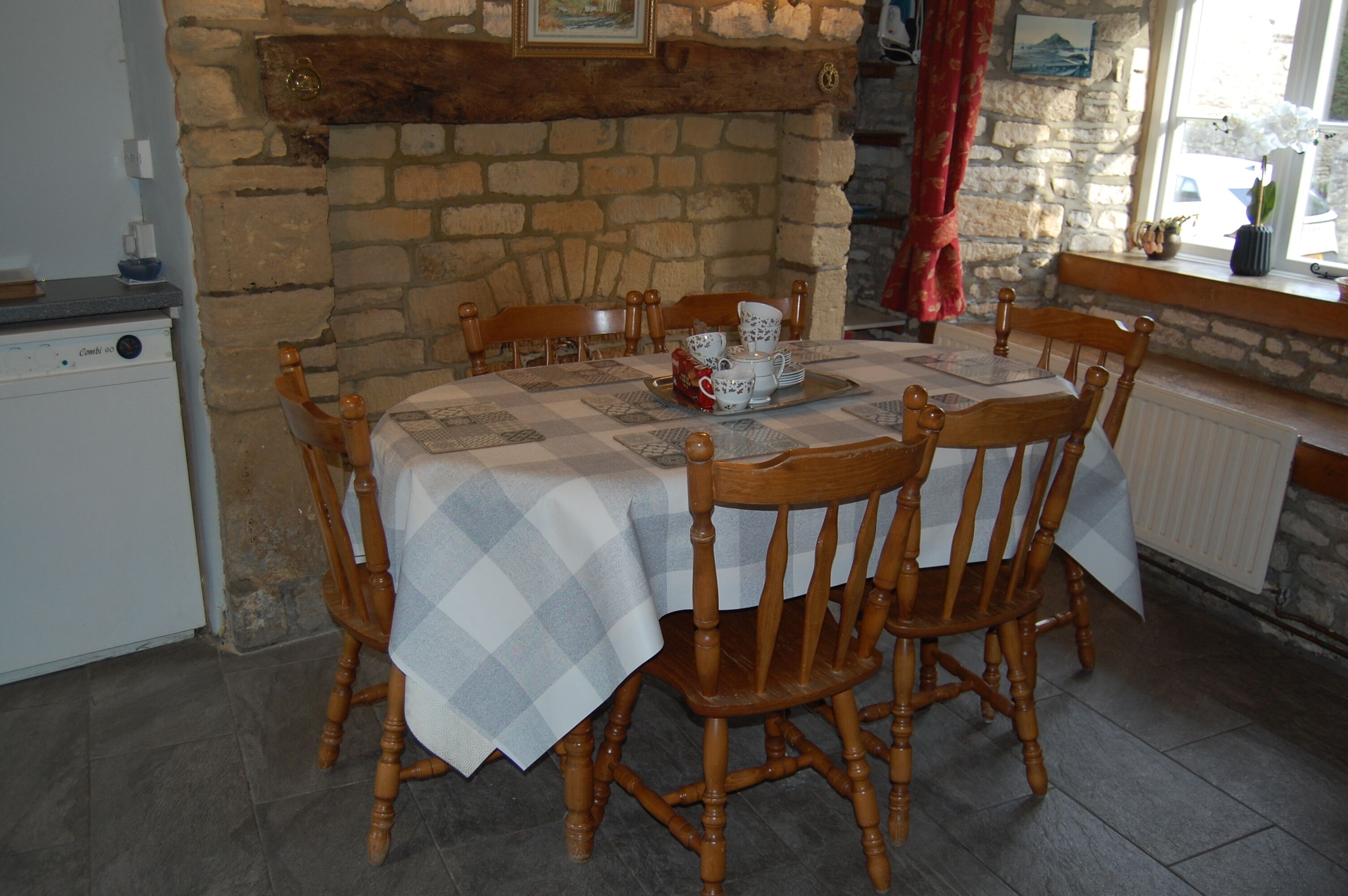 A dining area with a stone fireplace, wooden chairs, a table with a checkered tablecloth, and a window with a view of the outside.