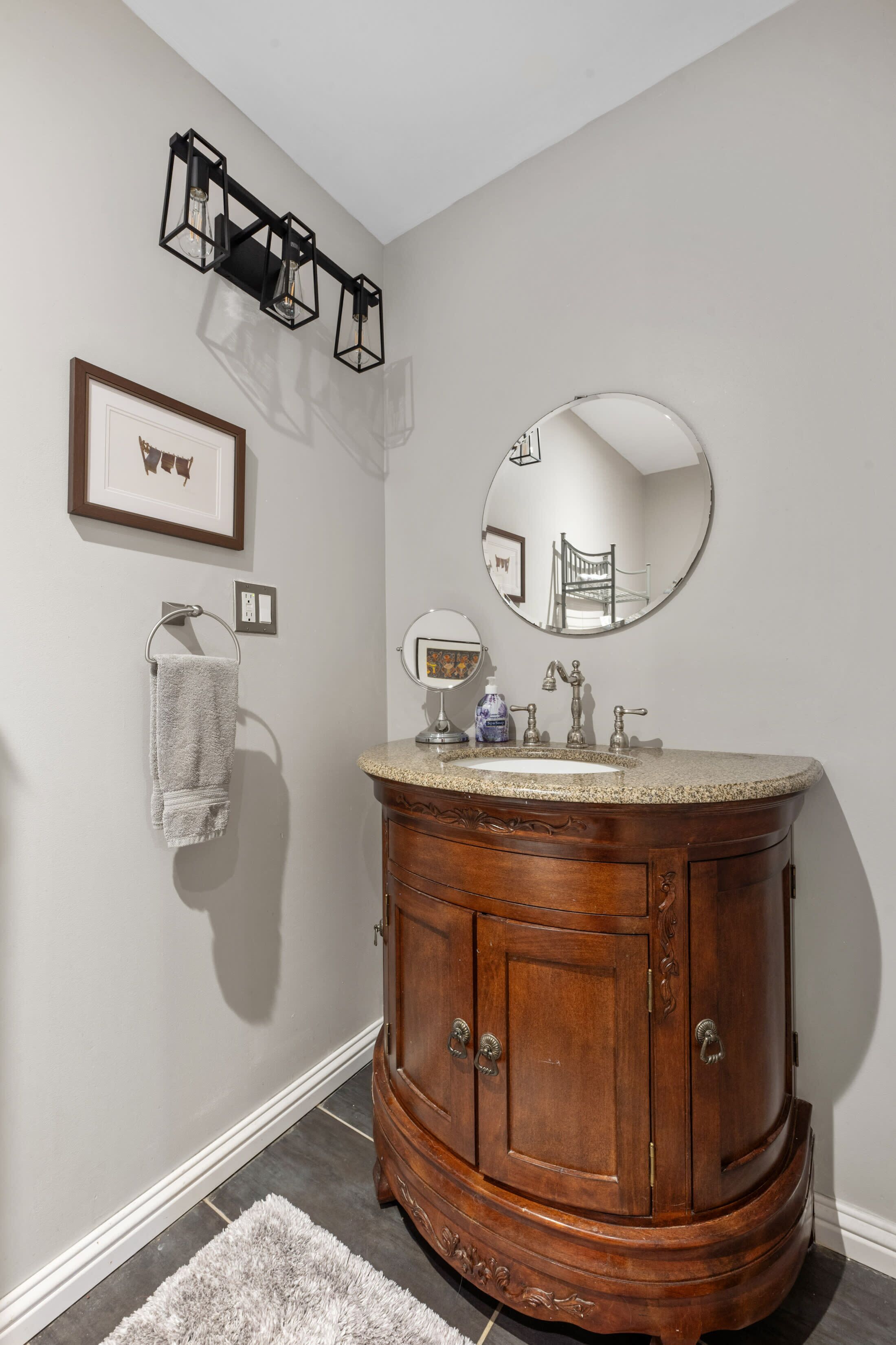 Un baño con tocador de madera, espejo redondo, cuadro enmarcado y perchero.