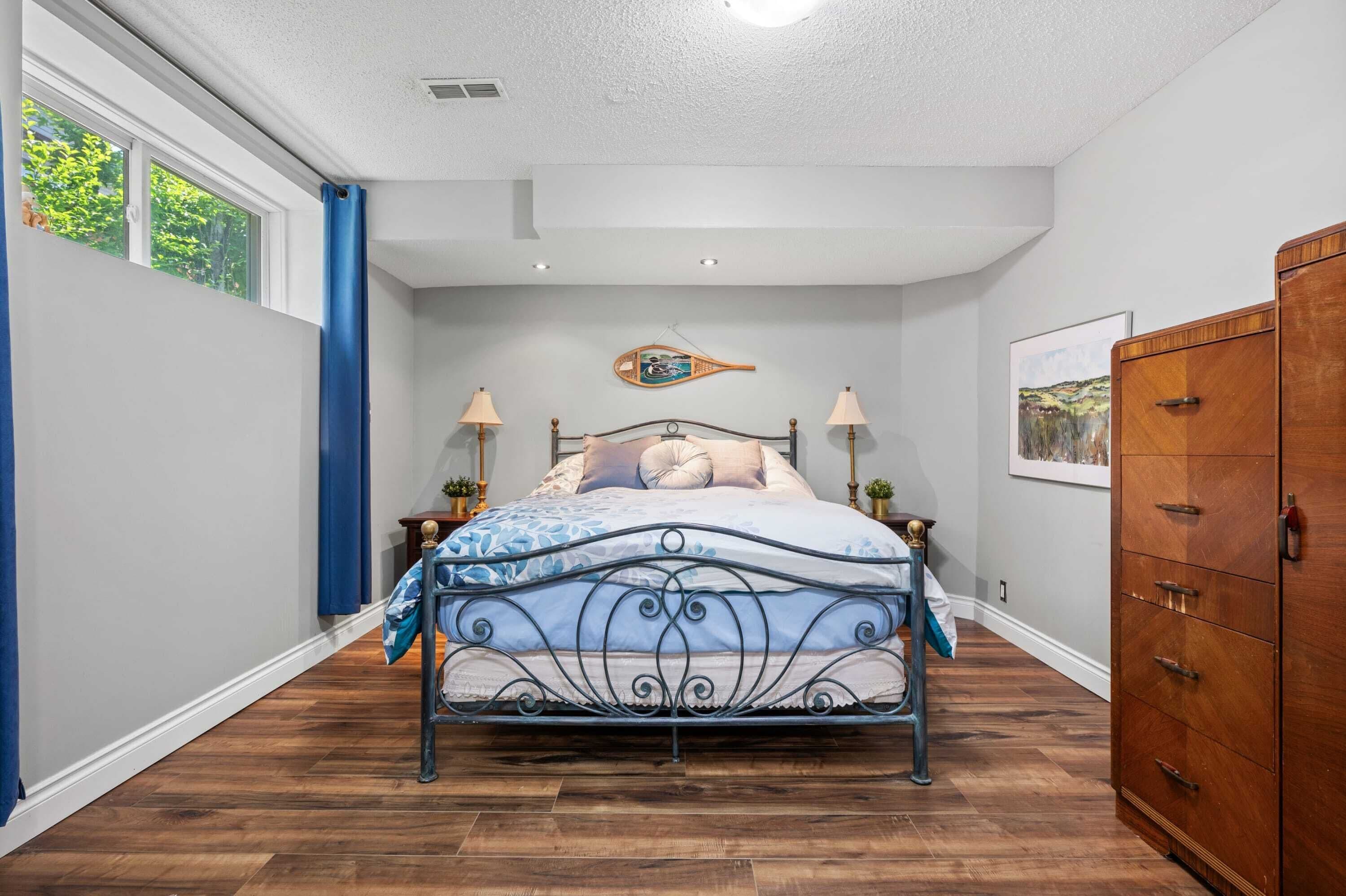 Een slaapkamer met een metalen bedframe, een houten dressoir en een raam met blauwe gordijnen.