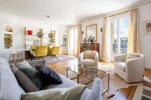 Una sala de estar con sofá, sillones, una mesa de centro, comedor con sillas, chimenea y ventanas amplias con cortinas.