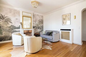 Uma sala de estar com lareira, um sofá, uma poltrona branca, um espelho e um mural na parede.