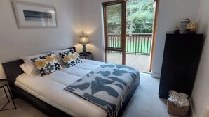 Ein Schlafzimmer mit einem Bett, einem Nachttisch, einer Lampe, einem Fenster mit Blick auf einen Garten und einer Tür.