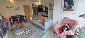 A living room with a sofa, a dining table with chairs, a pink armchair, and a kitchen in the background.