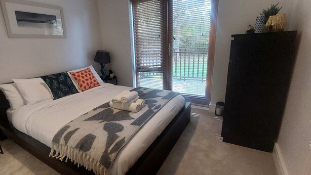 Ein Schlafzimmer mit einem Bett, einem Nachttisch, einer Lampe, einem Fenster mit Jalousien und einem dunkelbraunen Kleiderschrank.