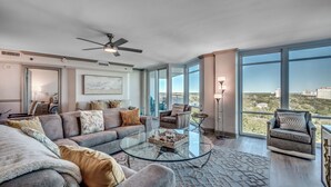 A spacious living room with a glass coffee table, grey sofas, and a ceiling fan. - ⛱️Ocean View Retreat with Private Balcony | Horizon at 77th - Unit 812🌊 (Myrtle Beach)