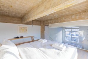 A bedroom with a large bed, a wooden bench, a framed picture, and a window with a view of buildings.