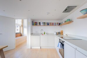 A modern kitchen with white cabinetry, a built-in bookshelf, and a window overlooking a brick wall. - Chic 1BD Warehouse Conversion, Hackney (Greater London)