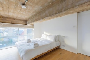 A bedroom with a large window, a bed with white linens, and a wooden ceiling.