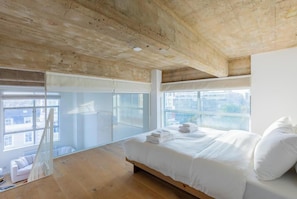 A bedroom with a wooden ceiling, large windows, a bed with white linens, and a view of the city.