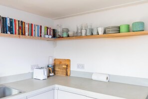 A kitchen with a countertop, a toaster, a blender, a cutting board, and a shelf with books and dishes.