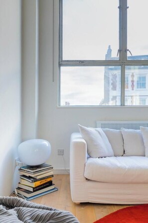A room with a white sofa, a stack of books, a white lamp, and a window with a view of buildings. - Chic 1BD Warehouse Conversion, Hackney (Greater London)