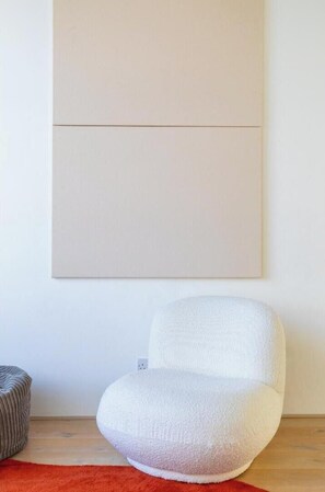 A room with a white chair, a red rug, and a large beige artwork on the wall.