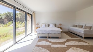 Een moderne slaapkamer met een groot bed, een kleinere bank en uitzicht op een tuin door raamwanden.