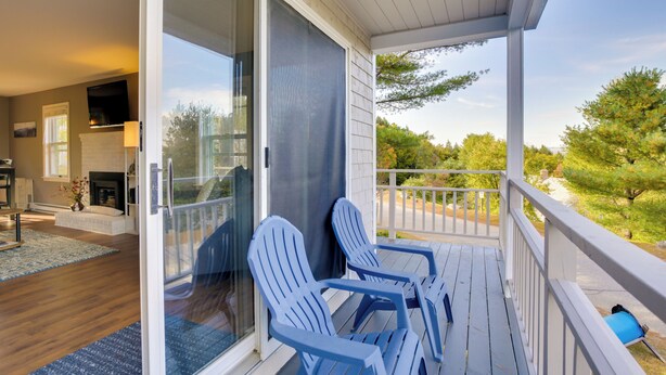 Eine überdachte Veranda mit zwei Adirondack-Stühlen, eine Glastür zu einem Wohnbereich mit Kamin und Blick auf Bäume und Himmel.