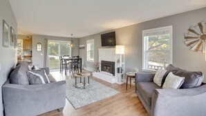 Ein modernes Wohnzimmer mit Kamin, grauen Sofas, einem Essbereich und großen Fenstern.