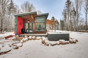 Une cabane moderne avec une façade en bois, de grandes fenêtres et un jacuzzi, située dans un cadre forestier enneigé.