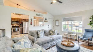 Una sala de estar moderna con un sofá seccional gris, un sillón azul y una mesa de centro de madera. Se ve una cocina al fondo con electrodomésticos de acero inoxidable.