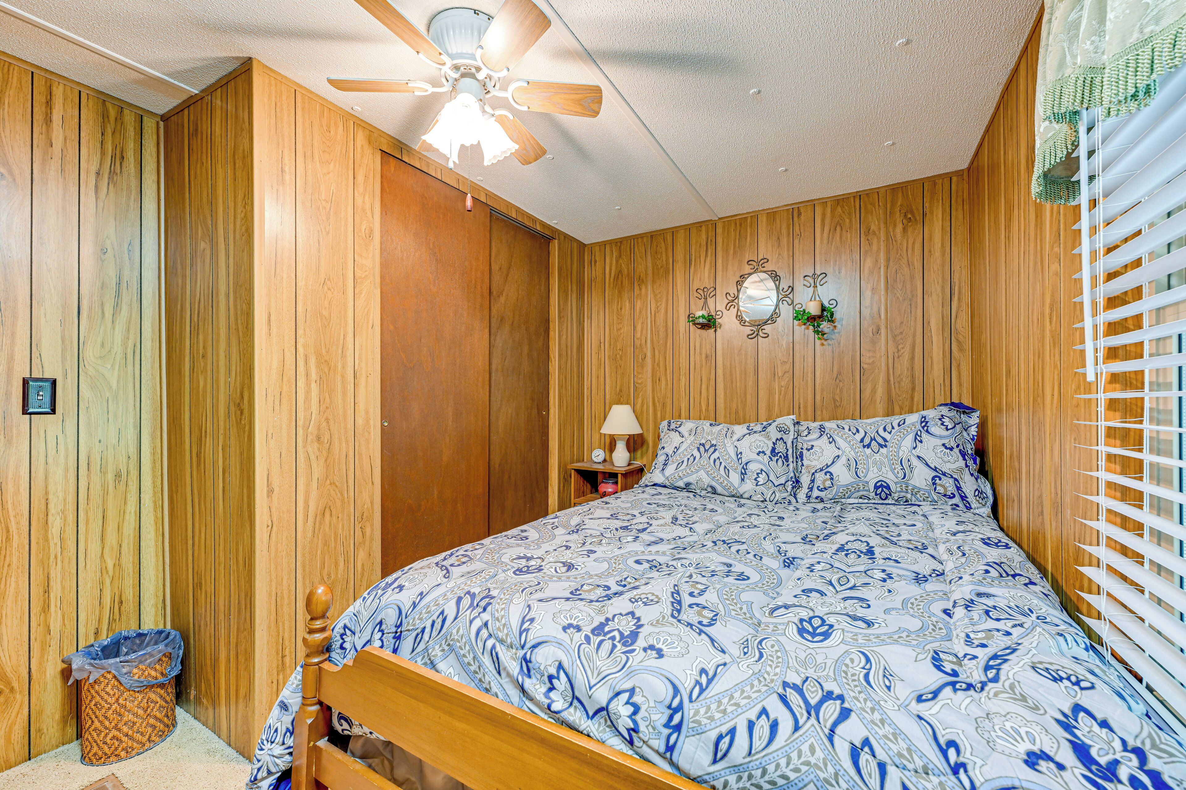 A bedroom with wooden paneling, a bed with a patterned bedspread, a ceiling fan, a nightstand with a lamp, and a window with blinds.