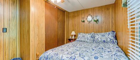 A bedroom with wooden paneling, a bed with a patterned bedspread, a ceiling fan, a nightstand with a lamp, and a window with blinds.