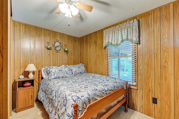 A bedroom with wooden paneling, a bed with a patterned bedspread, a nightstand with a lamp, and a window with blinds.