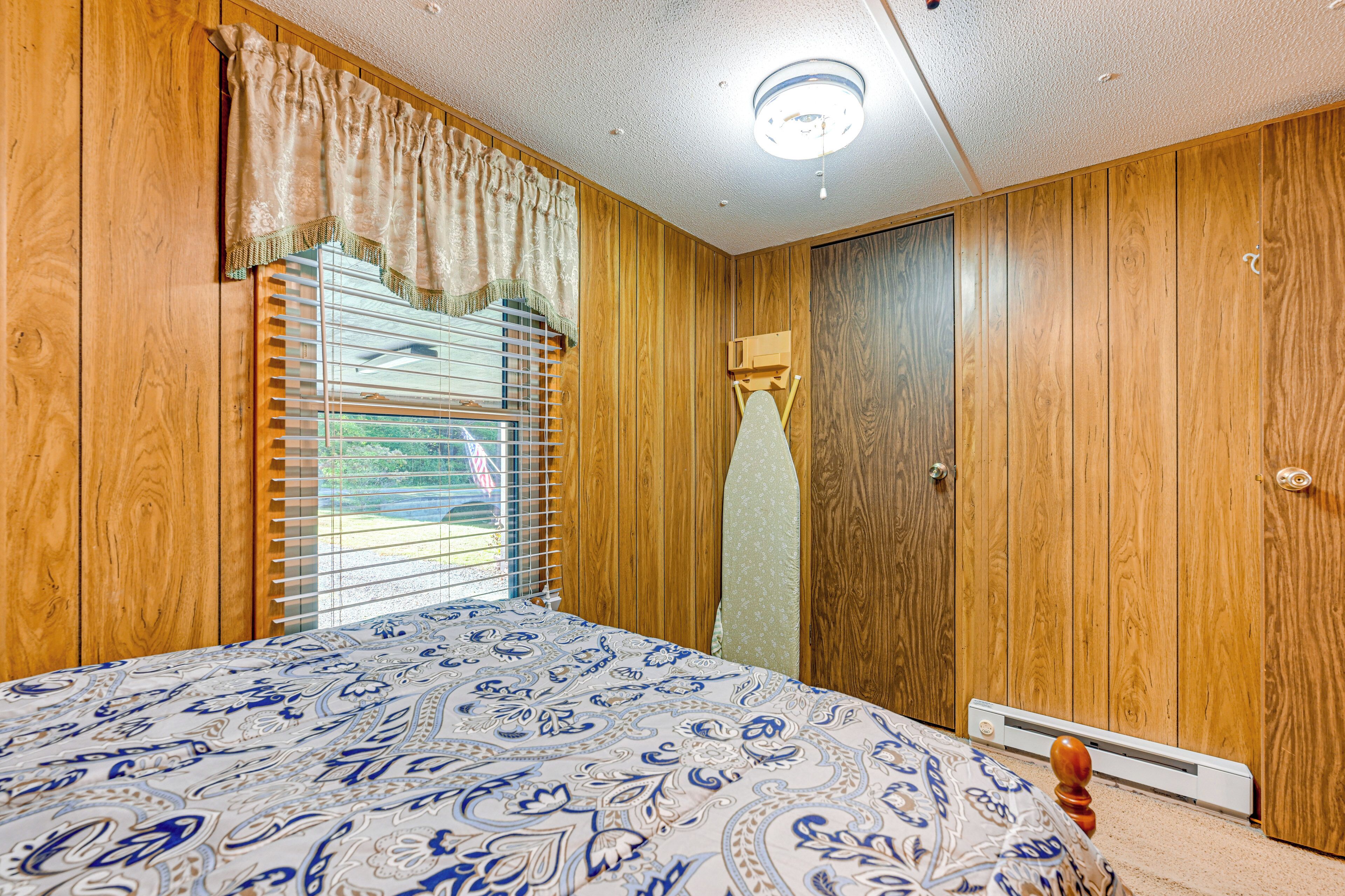 A bedroom with wooden paneling, a patterned bedspread, a window with blinds, and a ceiling light fixture.