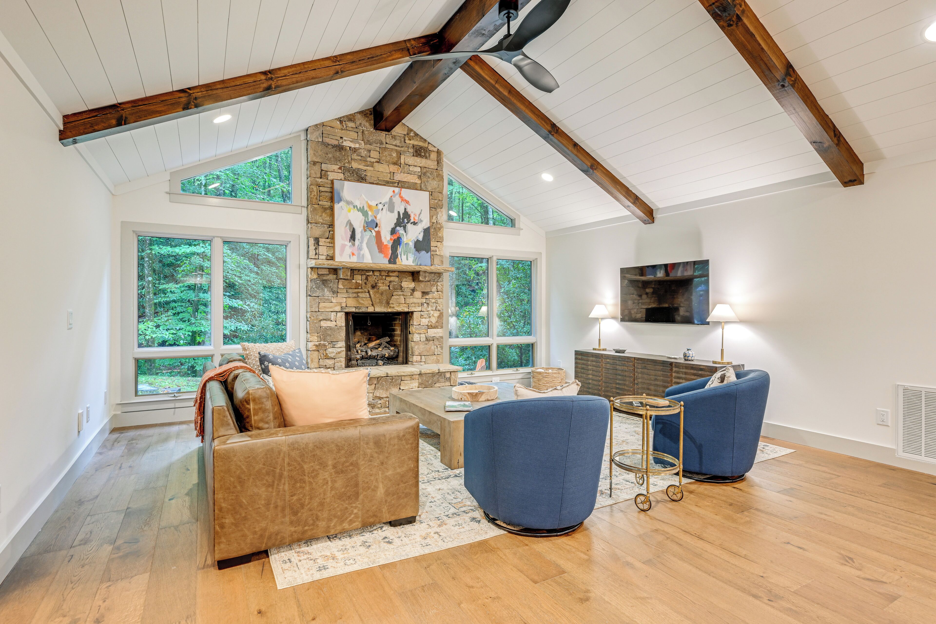 A spacious living room with a stone fireplace, large windows, and a high ceiling with exposed beams.