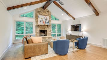 A spacious living room with a stone fireplace, large windows, and a high ceiling with exposed beams.
