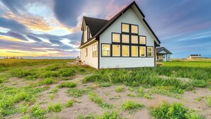 Une maison de plain-pied avec un toit à pignon et de grandes fenêtres, entourée d’un champ verdoyant et sous un ciel dégagé.