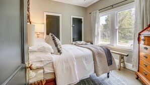 Een slaapkamer met een bed, een houten dressoir, een bankje en een raam met gordijnen.