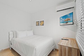 Ein Schlafzimmer mit einem Bett, einem kleinen Holfnachtkästchen, einer Wand-Klimaanlage und einem Bilderrahmen mit einer Strandansicht.