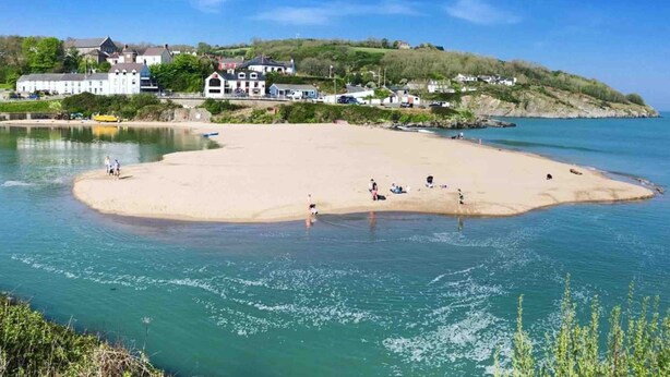 A coastal beach with a sandy shore, clear blue water, and buildings in the background.