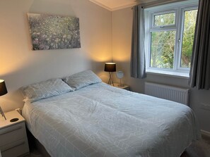 Ein Schlafzimmer mit Bett, Nachttisch, Lampe, einem Fenster mit Vorhängen und einem Bild an der Wand.