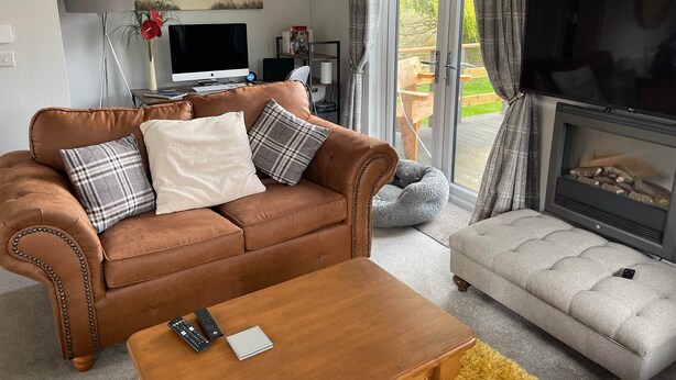 A living room with a brown leather sofa, a grey tufted ottoman, a wooden coffee table, a fireplace, a television, and a large window with curtains. - Holiday Let Tresaith - Clos Y Hendy Lodge (Tresaith)