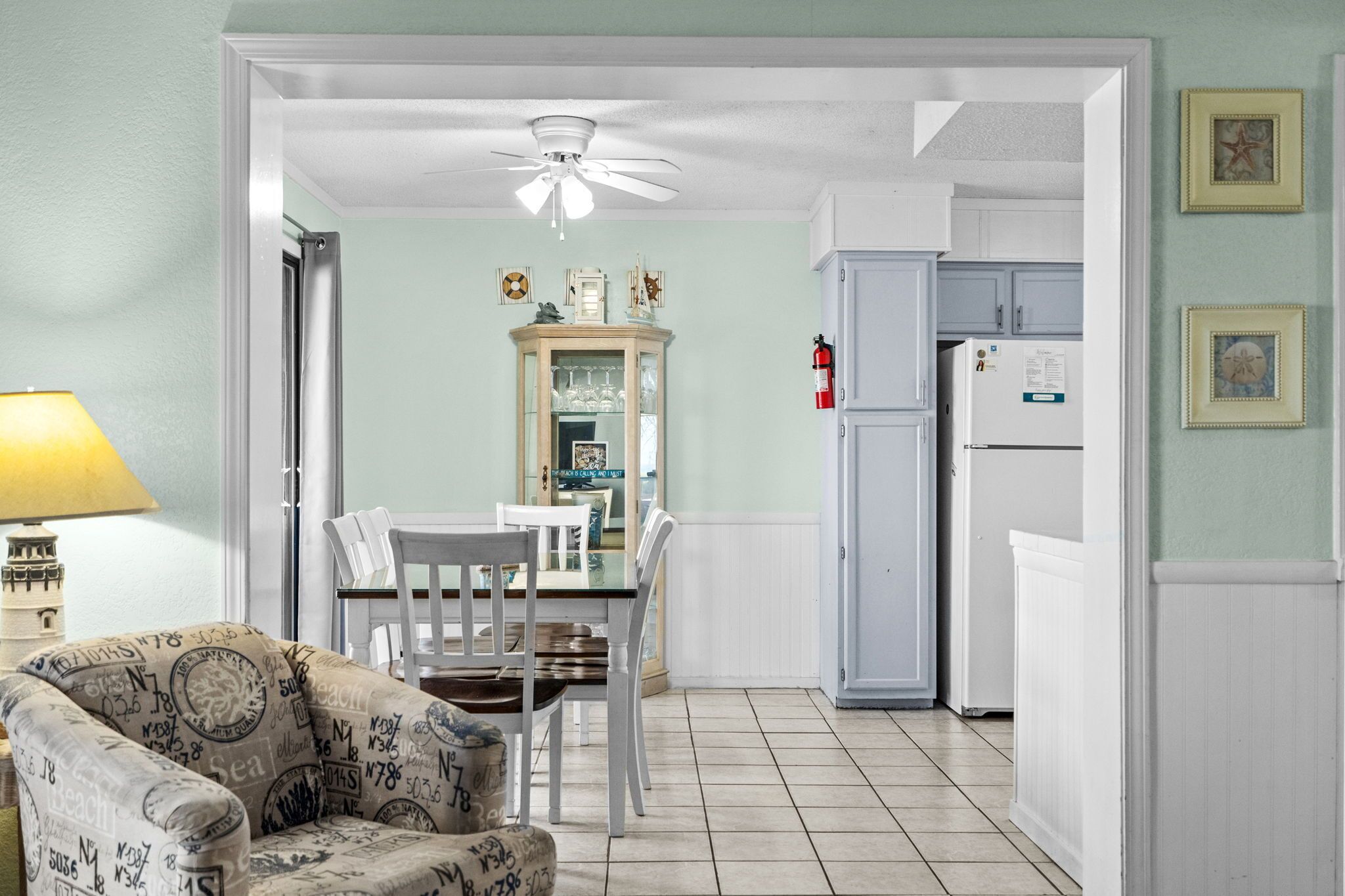Un coin repas comprenant une table et des chaises, un ventilateur de plafond, un meuble contenant de la verrerie, et un espace cuisine avec un réfrigérateur.