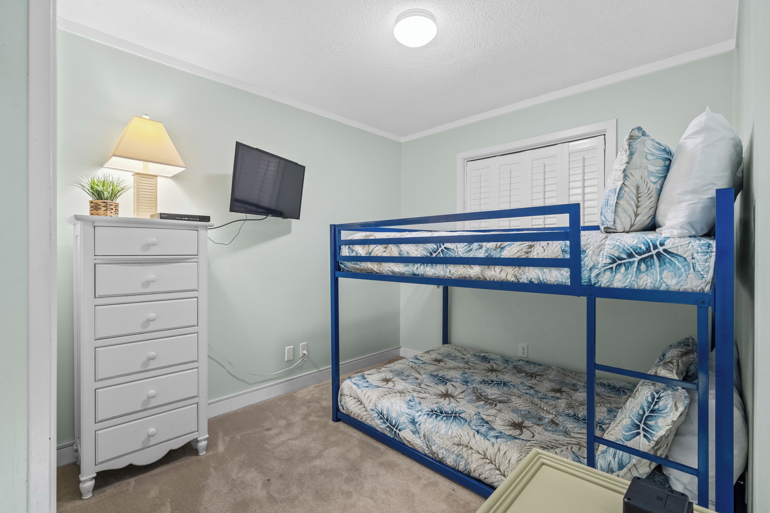 A bunk bed with a leaf-patterned mattress, a white dresser with drawers, a lamp, and a television on the wall.