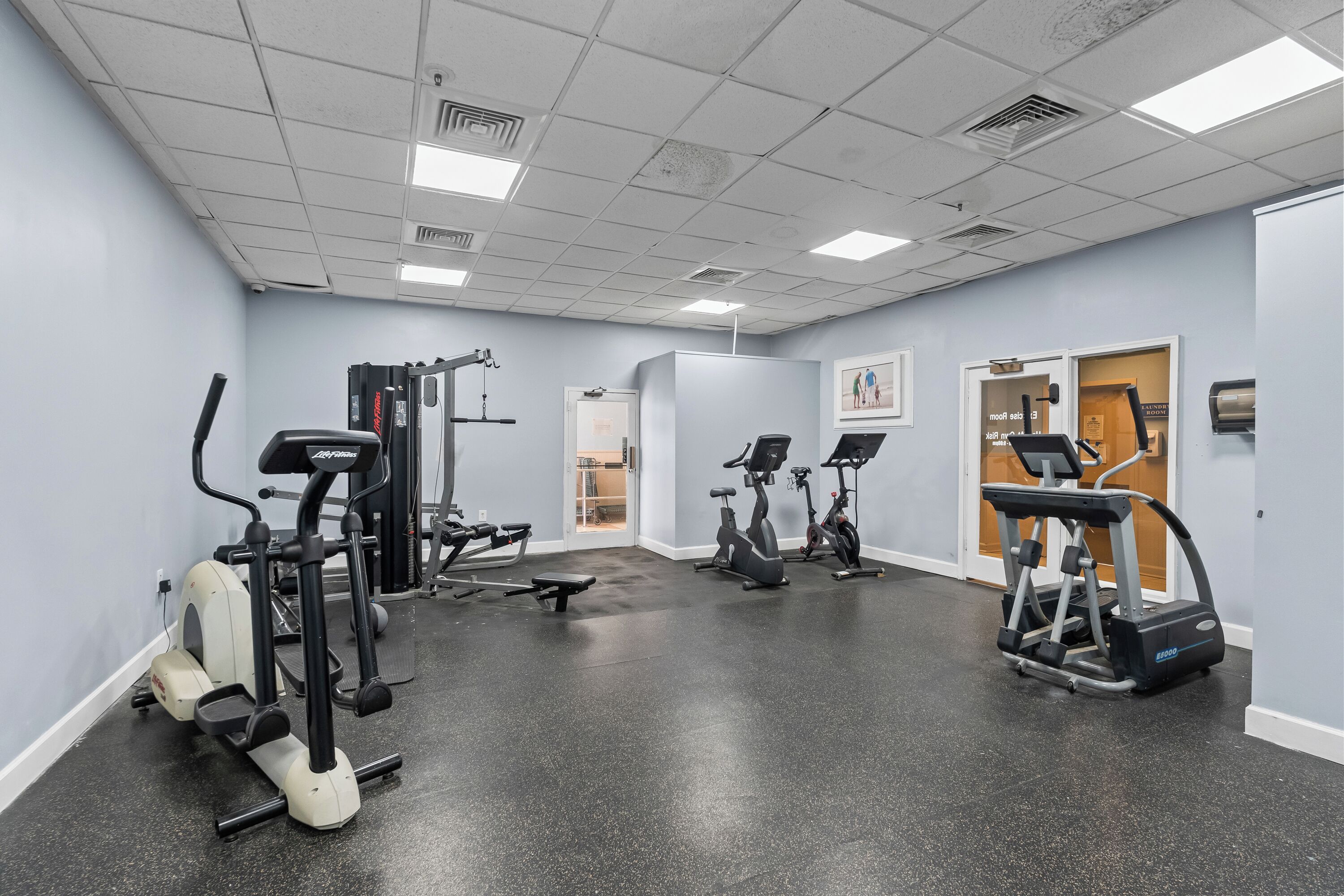 A gym with various exercise equipment, including treadmills and elliptical machines, and a door leading to another room.