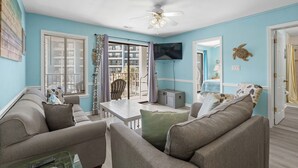Una sala de estar con ventilador de techo, televisor montado en la pared y vistas al balcón exterior.