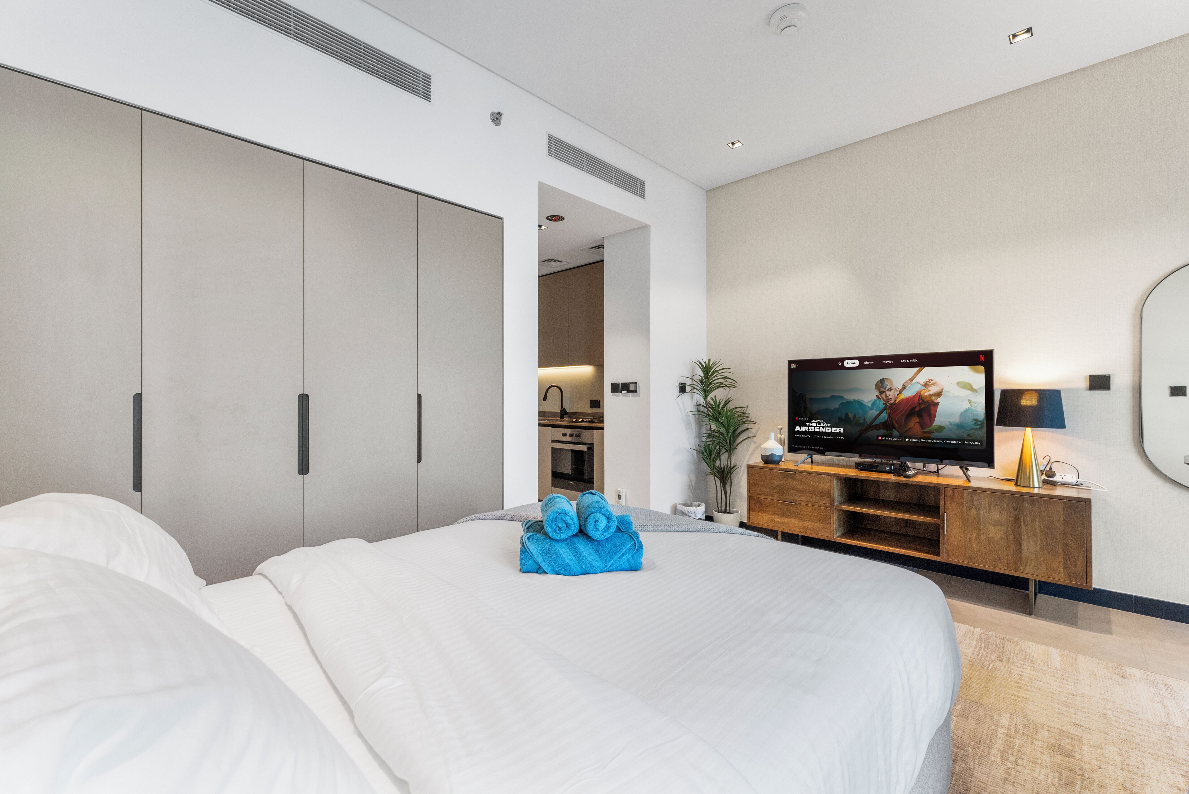 A modern hotel room with a large bed, a television, a wooden dresser, and a view into the kitchen area.
