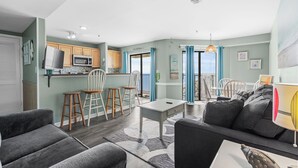 Een moderne woonkamer met een grijze bank, een eethoek met houten stoelen, een keuken met houten kasten en uitzicht op de zee door grote ramen.
